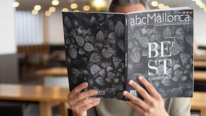 Una persona leyendo un libro llamado 'abcMallorca Best Addresses' en una mesa con cosas para el desayuno.