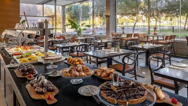 A buffet with various pastries and desserts, tables and chairs arranged for dining, and large windows offering a view of the outdoors. - Elba Almeria Business & Convention Hotel (Almeria)