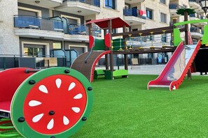 A playground with a slide and climbing structures, surrounded by a green artificial turf. - Apartamentos Nuriasol (Fuengirola)