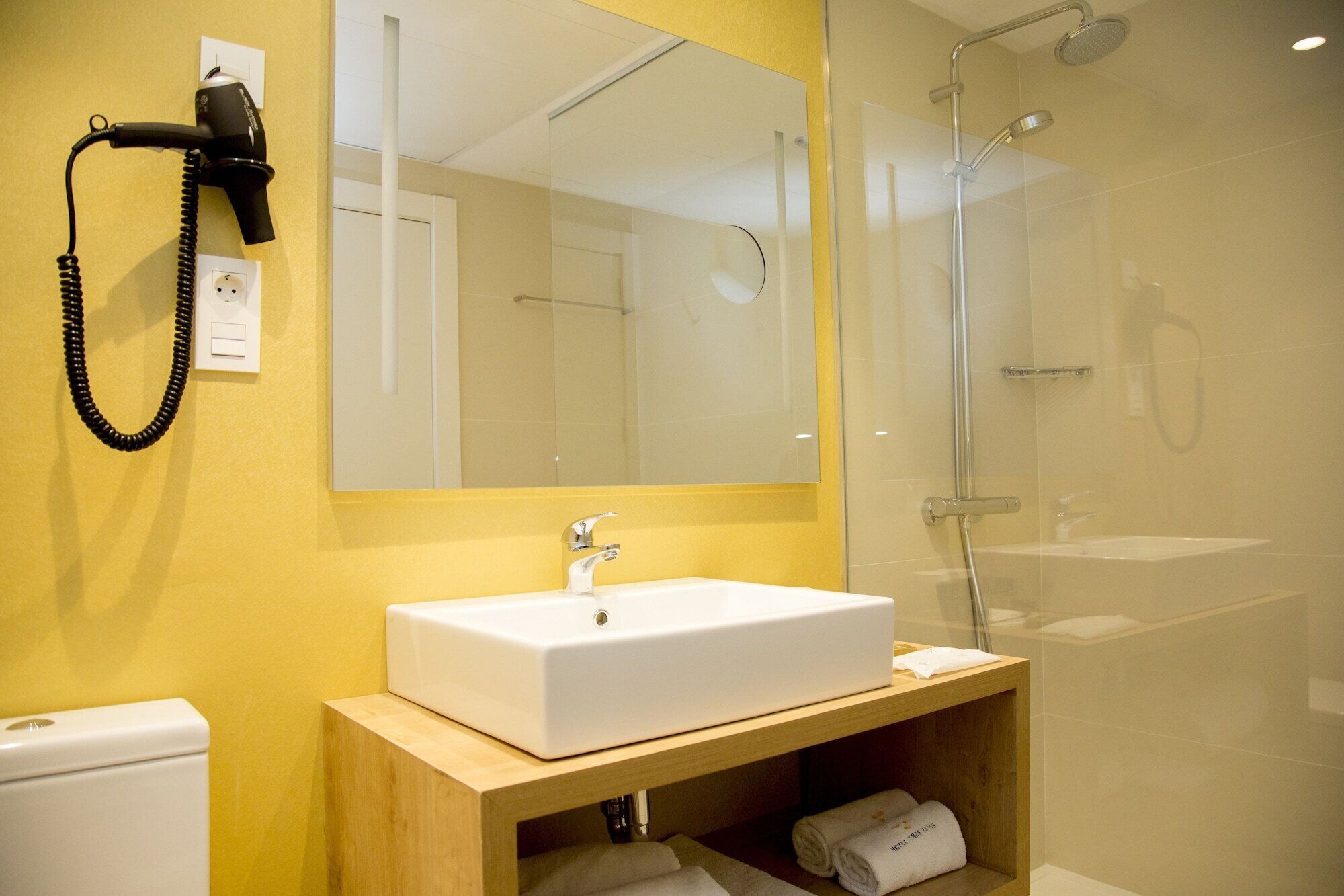 A modern bathroom with a white sink, a large mirror, a hairdryer, and a glass-enclosed shower.