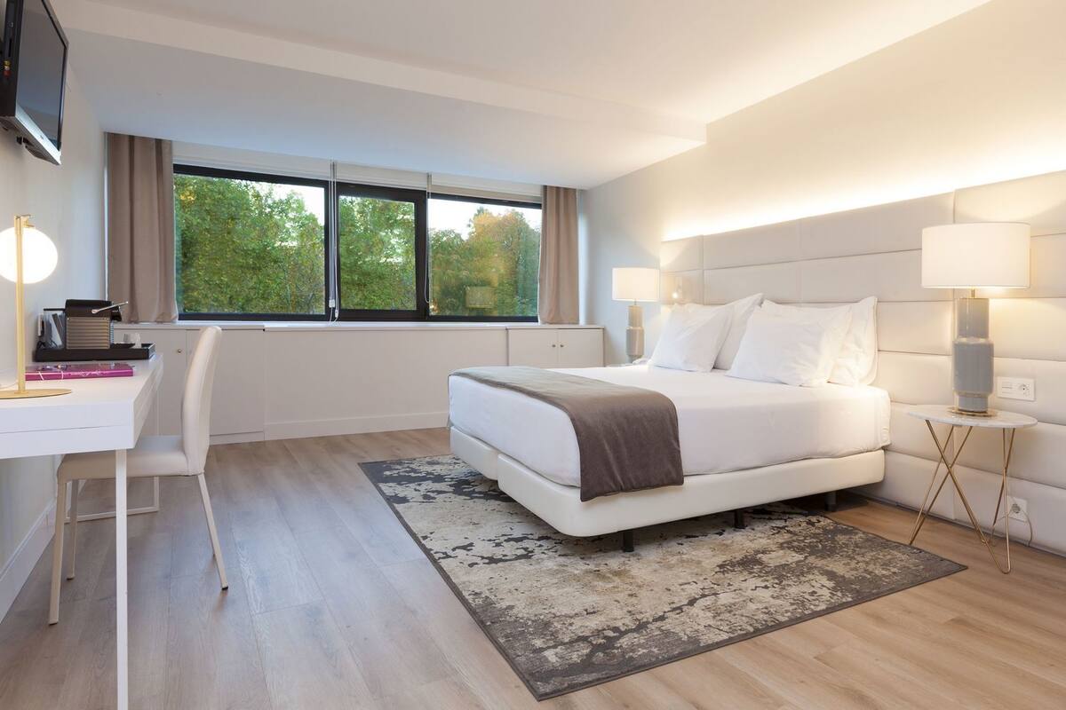 A modern hotel room with a bed, desk, and large windows.