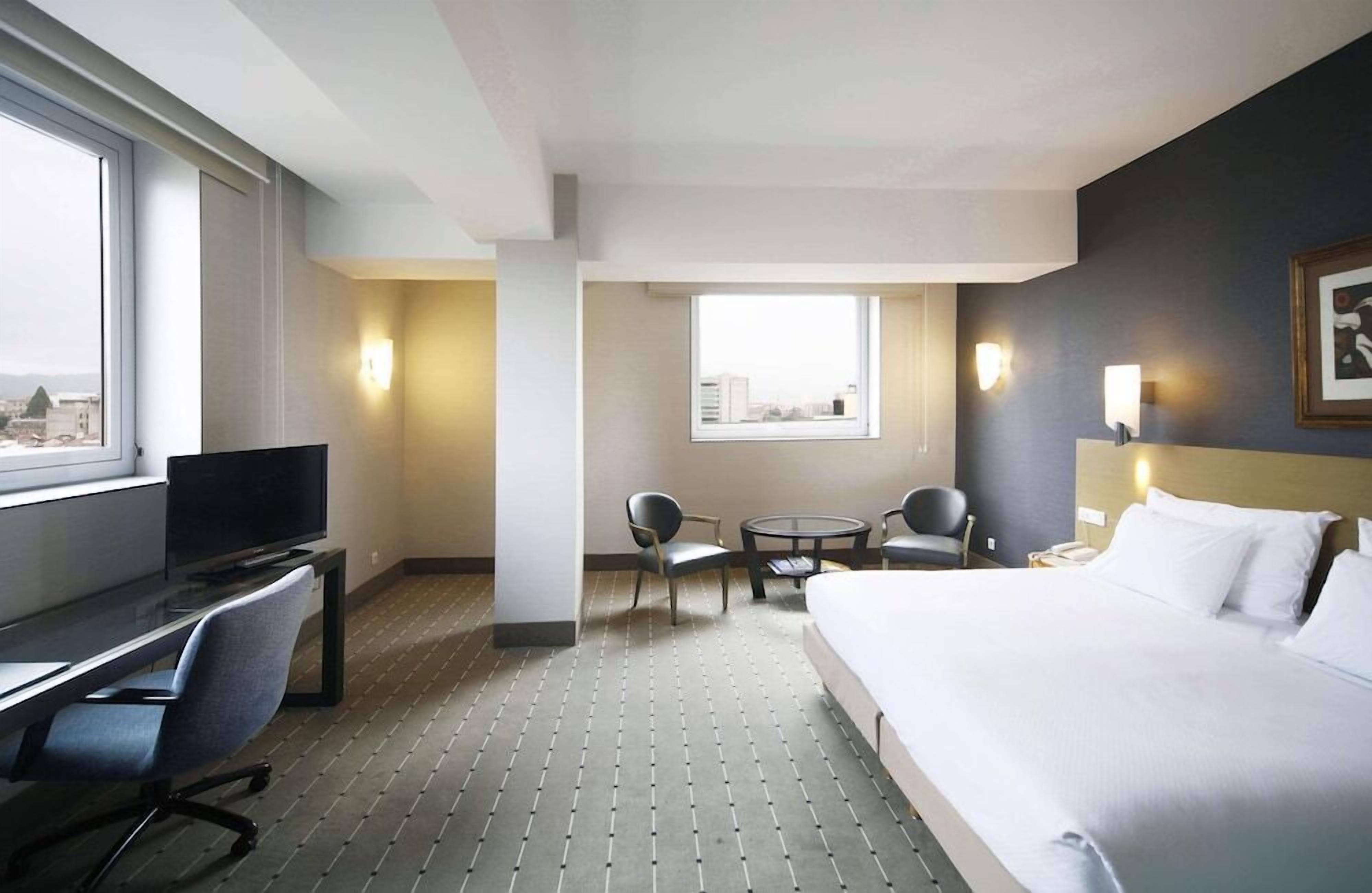Habitación de hotel con cama, escritorio, televisor, dos sillas, una mesita y ventana con vistas a la ciudad.