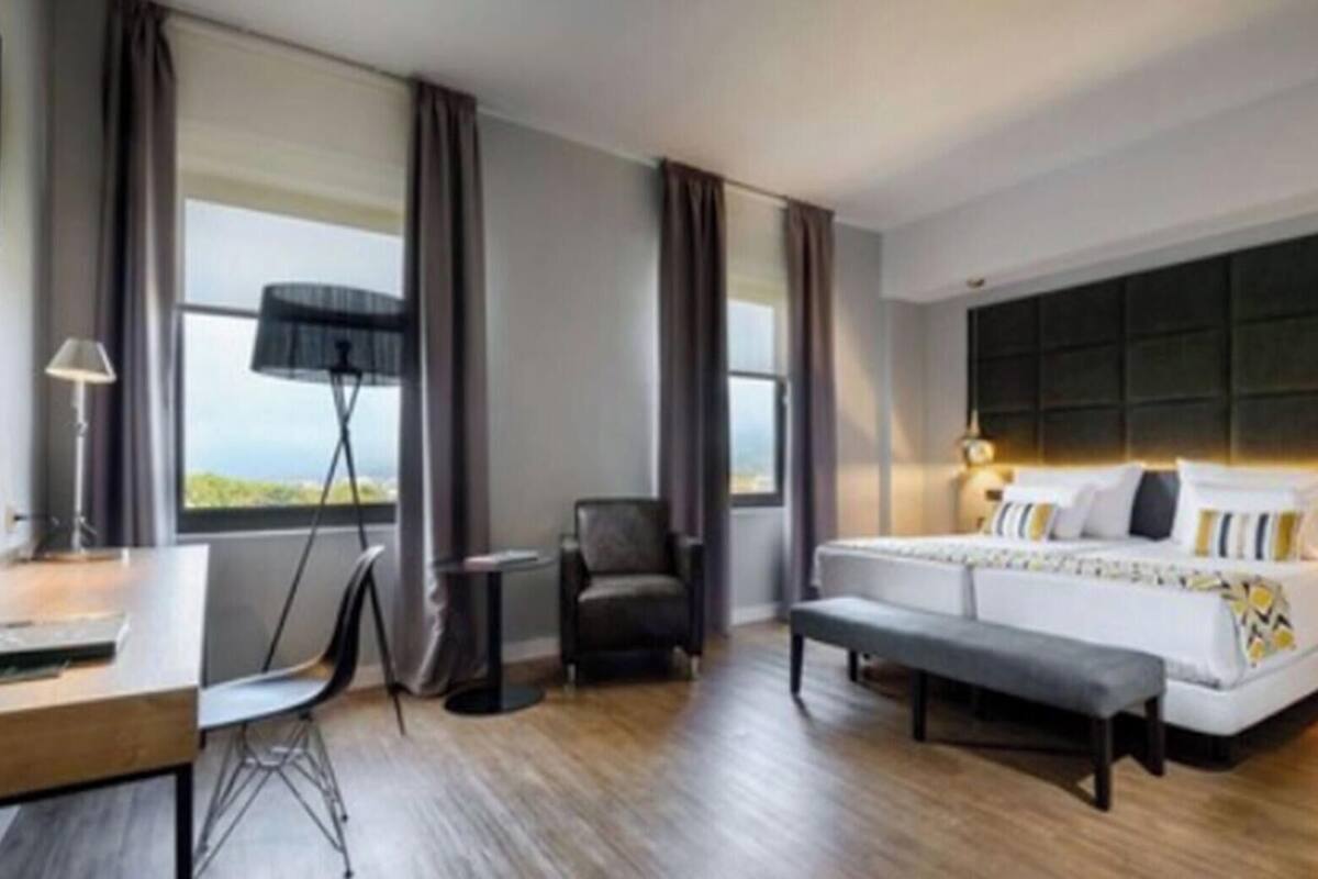 A modern hotel room with a large bed, a desk, a chair, and a view of the outdoors.