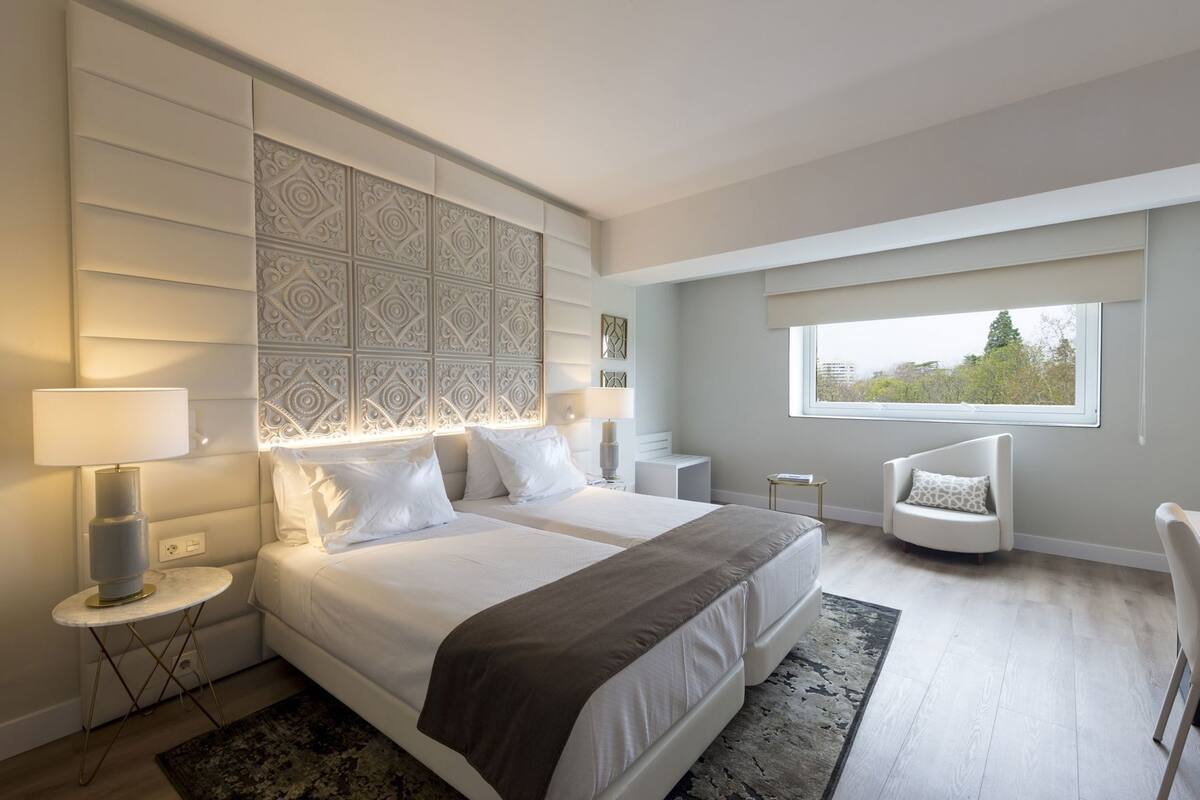 A modern hotel room with a large bed, bedside tables, a chair, a window, and a decorative headboard.