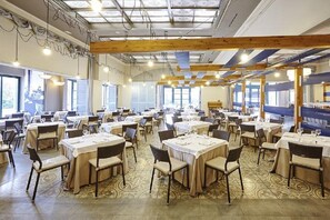 Un vaste espace dédié à la salle à manger, avec de nombreuses tables et chaises, de grandes fenêtres et un plafond haut avec des poutres apparentes.