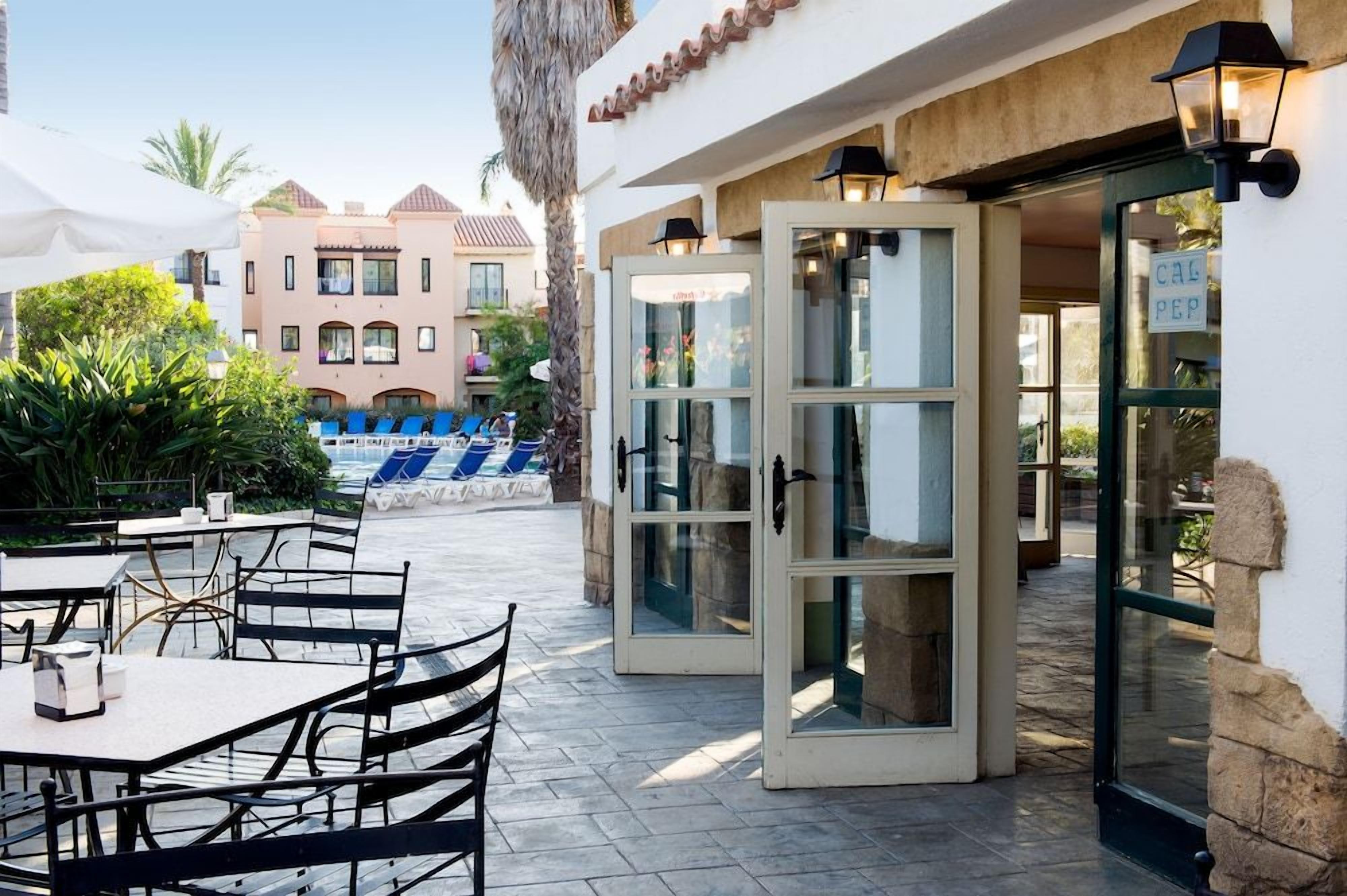 An outdoor patio with tables and chairs, a pool area, and a building entrance with glass doors.