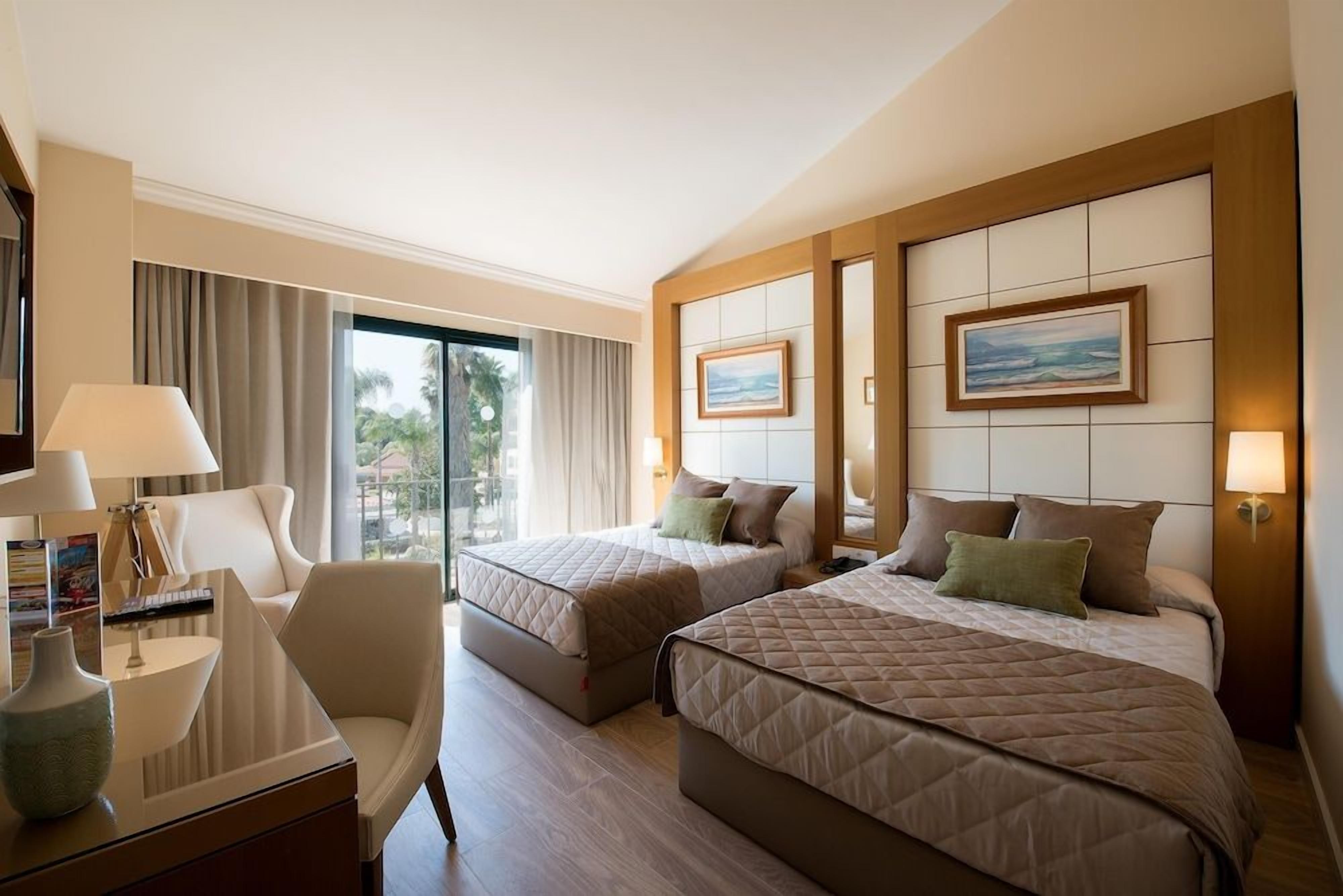 A hotel room with two beds, a desk, a chair, and a large window with curtains.