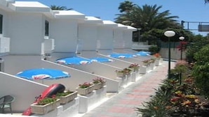 A row of white buildings with blue umbrellas, a pathway, and potted plants. - Niza Apartments (Mogan)