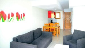 A living room with a grey sofa, a dining table with orange chairs, and a kitchen area in the background. - Niza Apartments (Mogan)