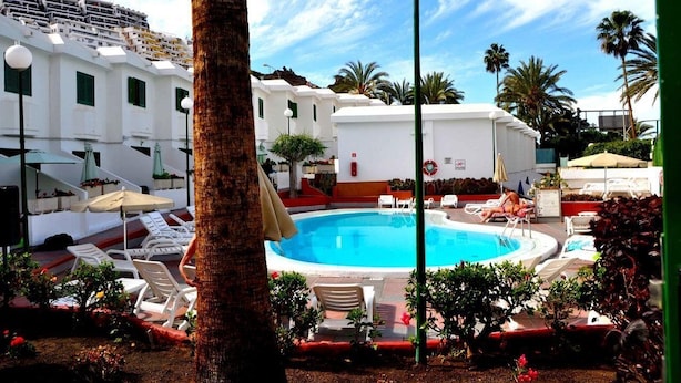 A pool area with lounge chairs and umbrellas, surrounded by white buildings and palm trees. - Niza Apartments (Mogan)
