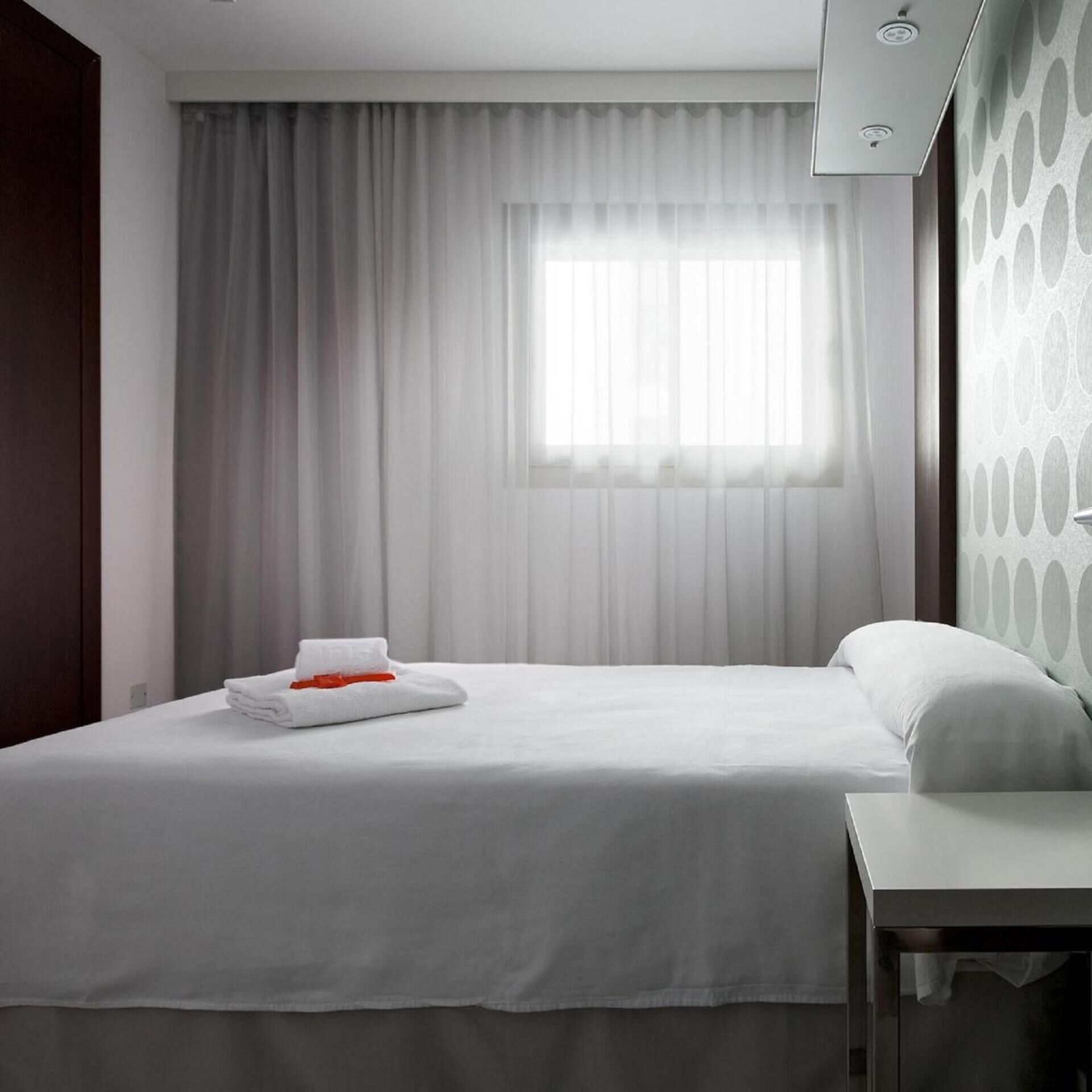 A hotel room with a bed, a nightstand, a window with curtains, and a wall with a circular pattern.