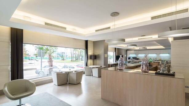 A modern hotel lobby with a reception desk, seating area, and large windows offering a view of the street. - Suittes Hotel (Algeciras)