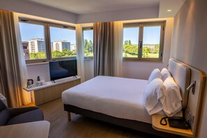 Une chambre d’hôtel équipée d’un lit, d’une télévision, d’un bureau avec un téléphone et de fenêtres donnant sur des immeubles et des arbres.