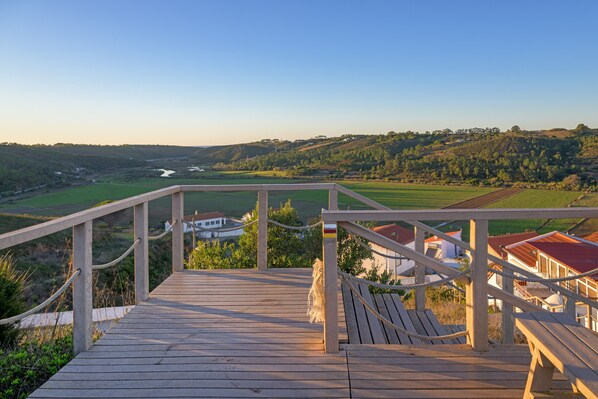 Terraza de madera con barandilla que da a un valle con casas y un río.