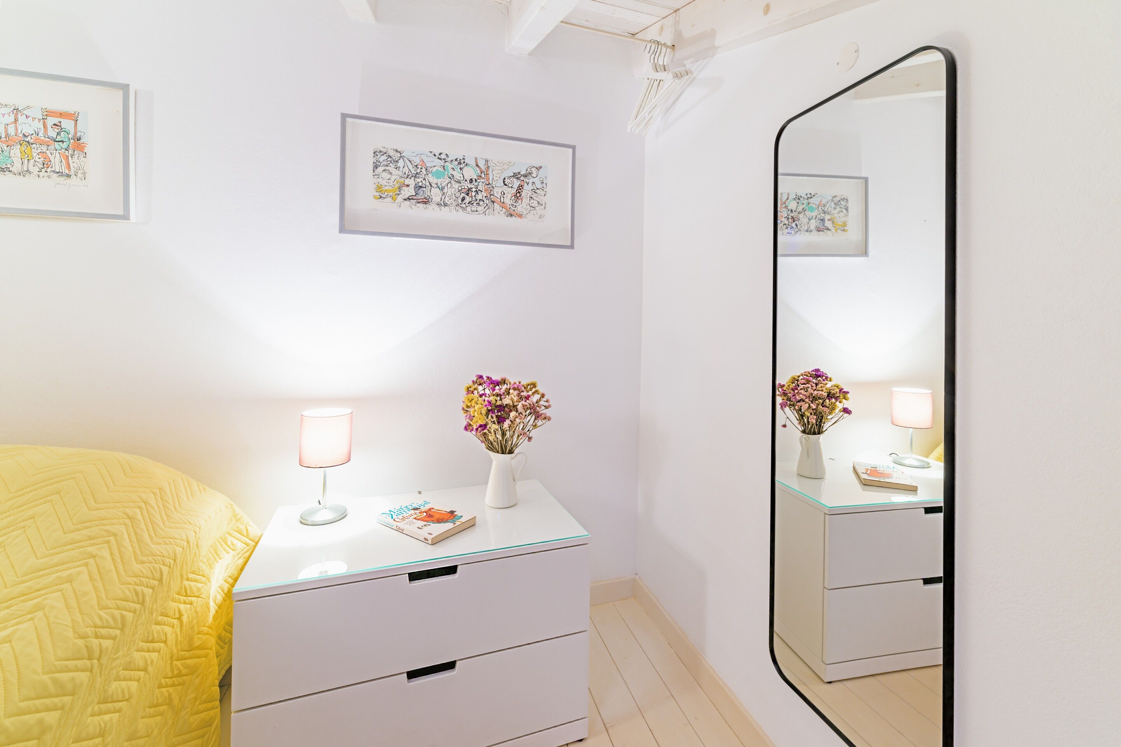 A bedroom with a bed, bedside table, mirror, and artwork on the wall.