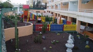 A courtyard with a children's play area, including colorful fences and a slide, surrounded by buildings. - Montemar Apartments (San Bartolomé de Tirajana)