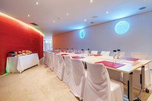 Una sala conferenze con un lungo tavolo, sedie bianche e una parete rossa.