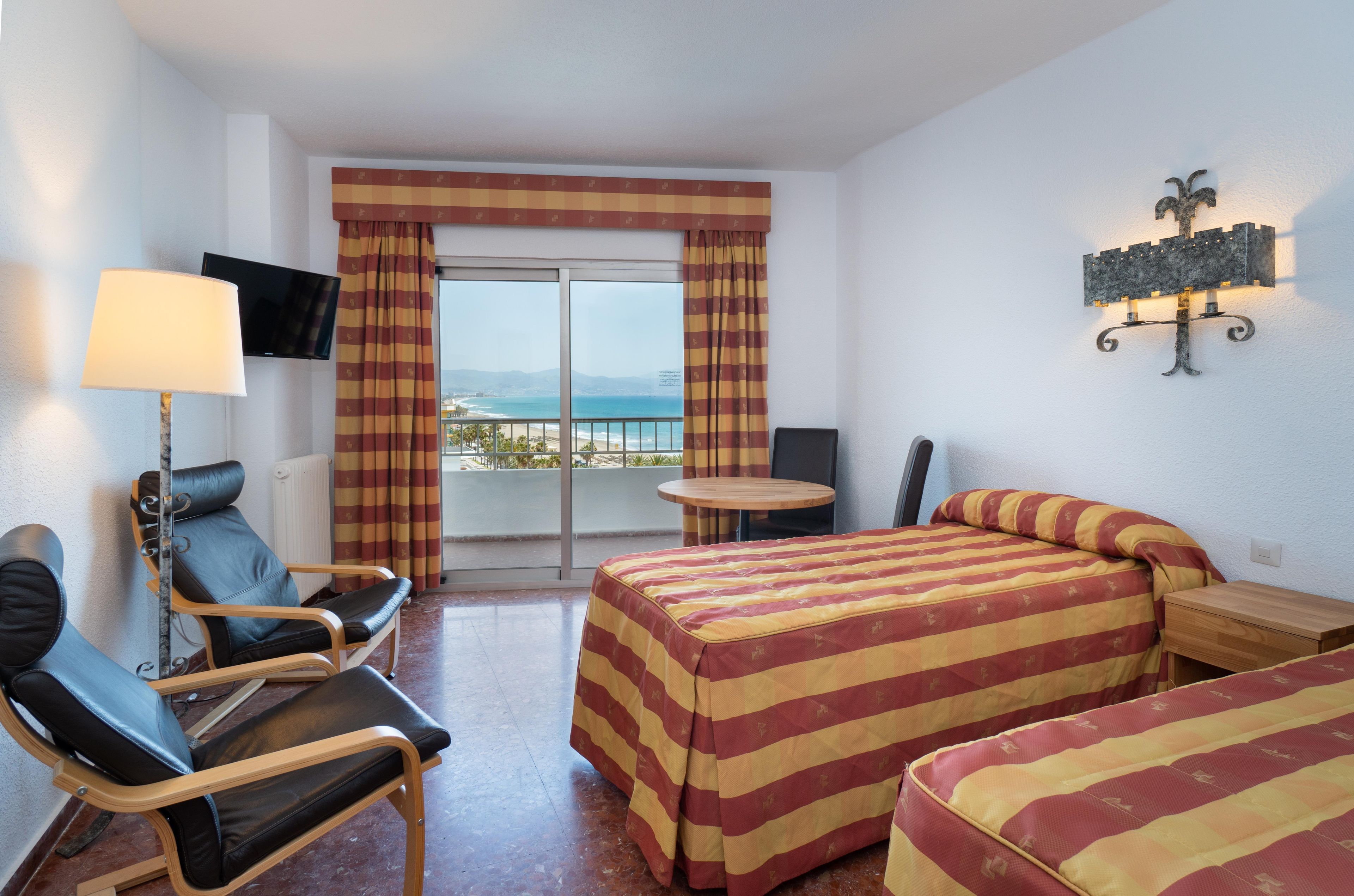 A hotel room with a bed, a chair, a table, a TV, and a view of the ocean.