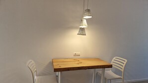A small dining area with a wooden table and two white chairs, a hanging light fixture above, and a white wall with a power outlet. - Apartamentos Costamar 2 (Formentera)
