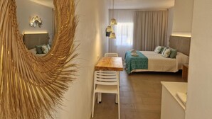 A hotel room with a large mirror, a bed with a green bedspread, a wooden table, white chairs, and a hanging light fixture. - Apartamentos Costamar 2 (Formentera)