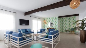 A modern living room with white furniture, a large TV, and a tropical wallpaper. - Apartamentos Costamar 2 (Formentera)