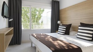 A modern hotel room with a large bed, a TV, and a desk. There is a window with curtains, and the room has a wooden headboard.