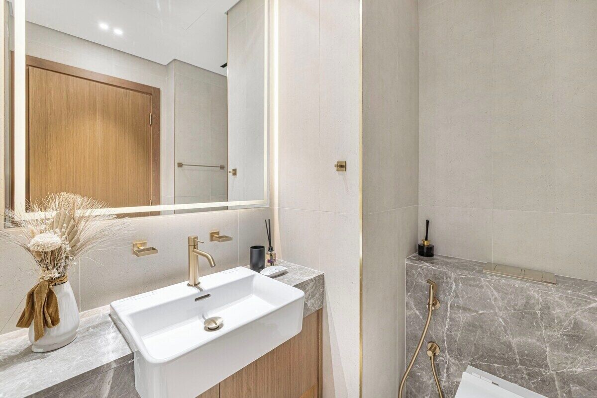 Un baño moderno con lavamanos blanco, detalles dorados y zona de ducha de mármol.