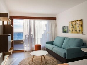 Een woonkamer met een bank, salontafel en televisie. Er is een balkon met uitzicht op zee.