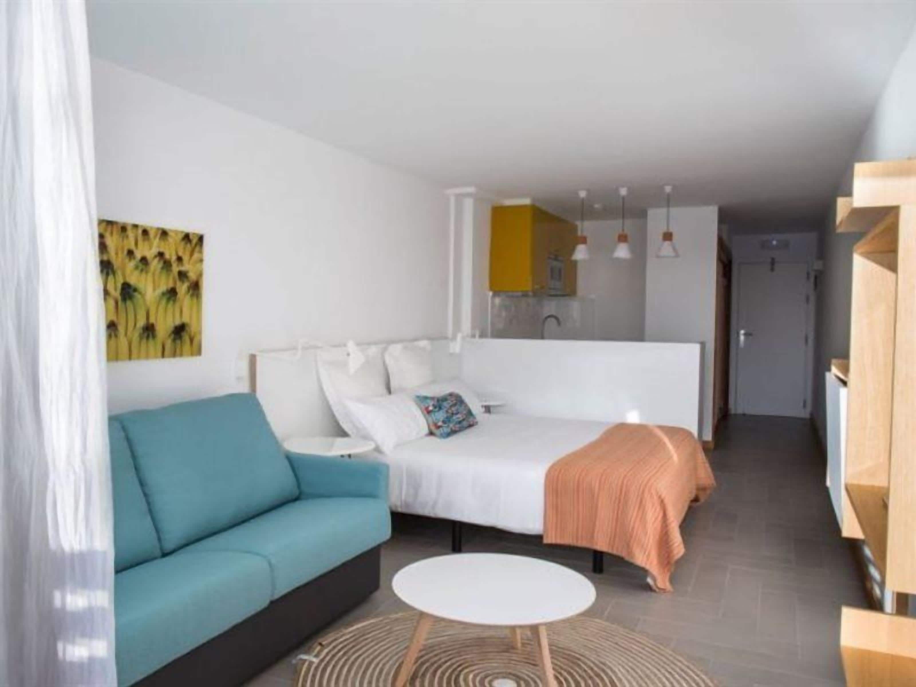 A modern bedroom with a bed, a sofa, a small round table, and a kitchen area in the background.