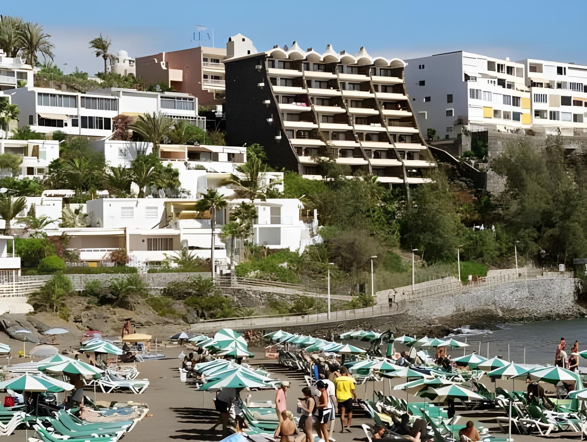 Une station balnéaire dotée d’immeubles à plusieurs étages, d’une plage aménagée avec des parasols et des chaises longues, et d’un rivage rocheux.