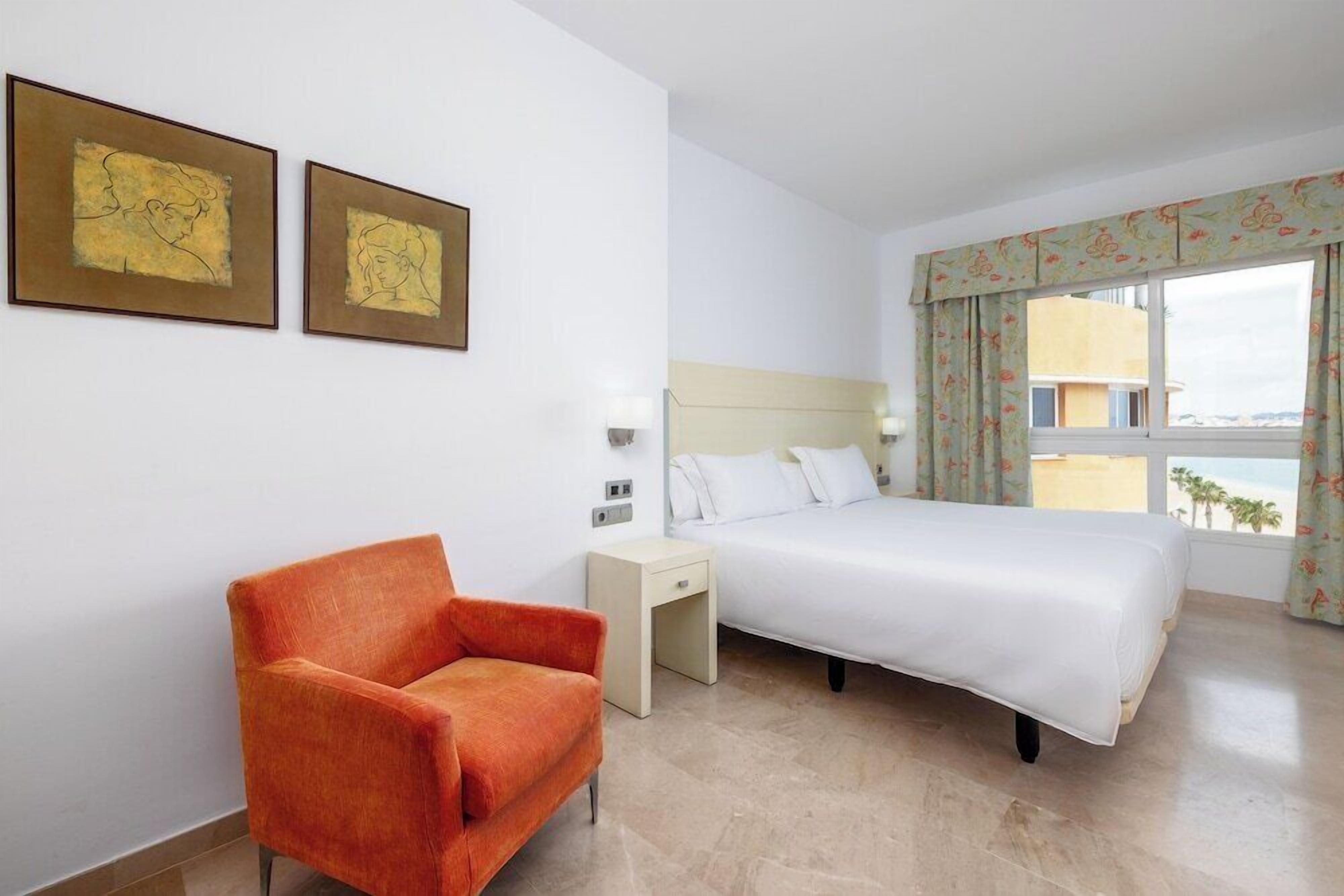 Een hotelkamer met een bed, een stoel, een nachtkastje en twee kunstwerken in lijsten aan de muur.