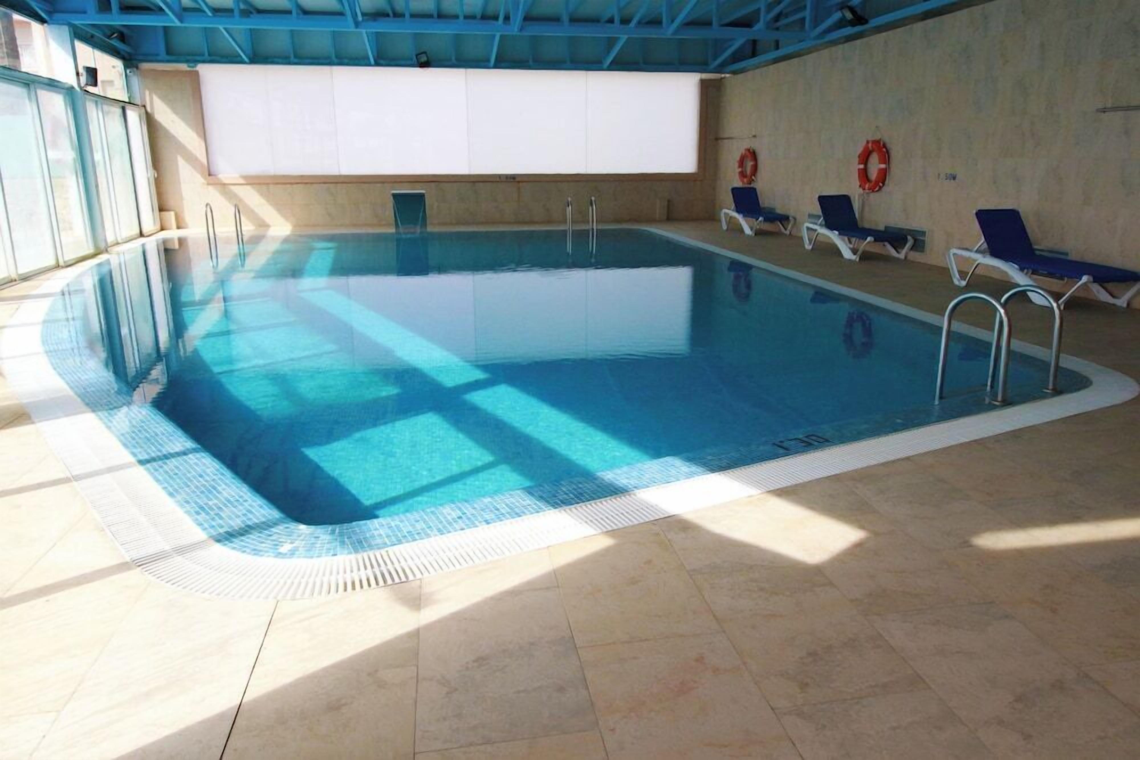 An indoor swimming pool with clear blue water, tiled floor, and blue lounge chairs.
