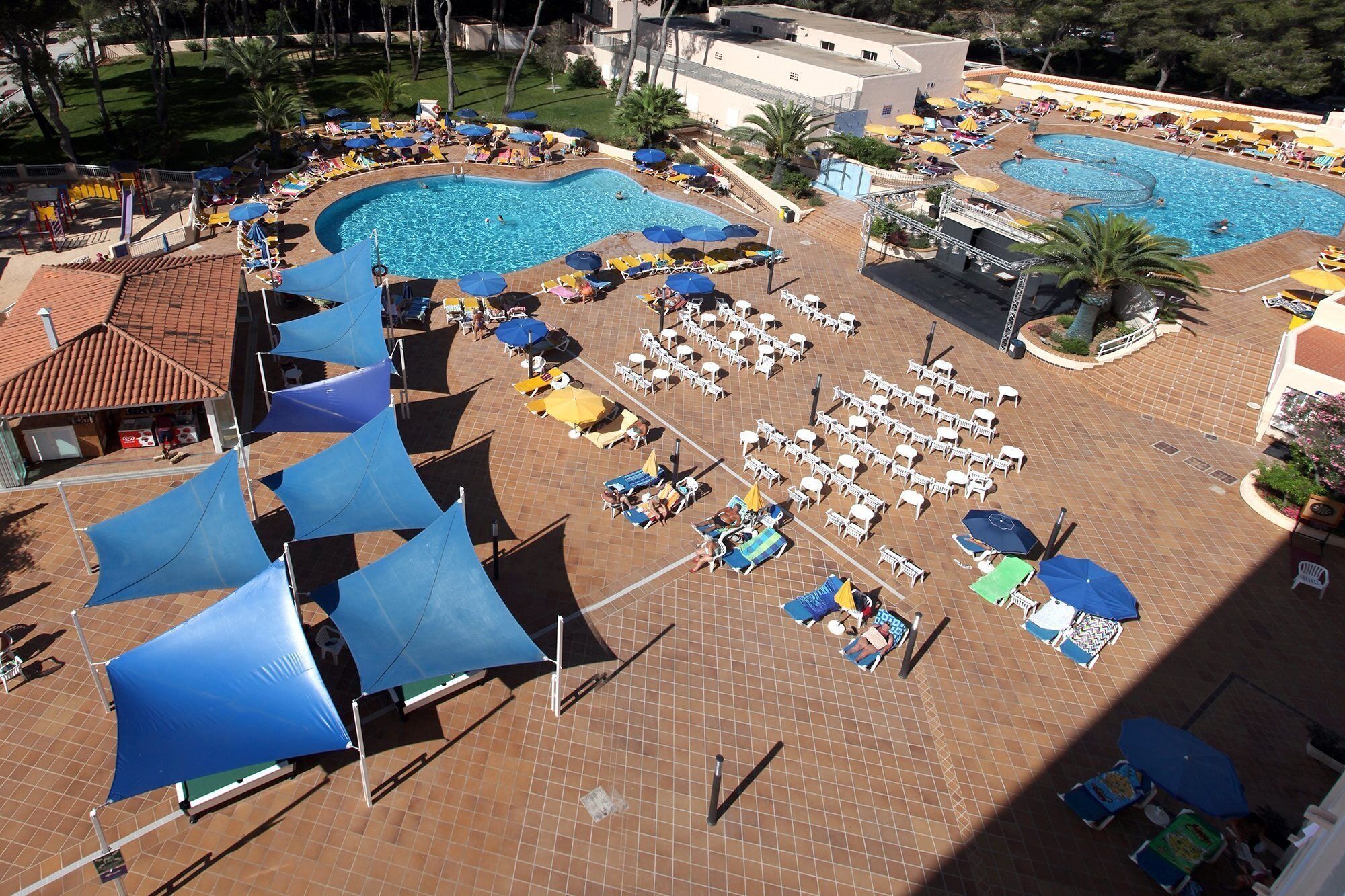 Zona de piscina con varios sillones de sol y sombrillas, rodeada de senderos de baldosas y un edificio con tejado de cerámica.