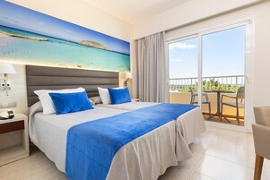 Habitación de hotel con una cama, una mesita de noche, una lámpara, un cuadro de una playa y un balcón con una mesa y sillas.