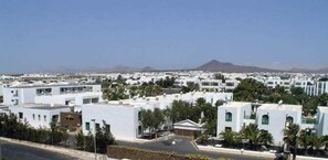 Zona residencial con edificios blancos, algunos con techos planos y otros con techos inclinados, organizados en un patrón de cuadrícula. Se observan palmeras y un cielo despejado.