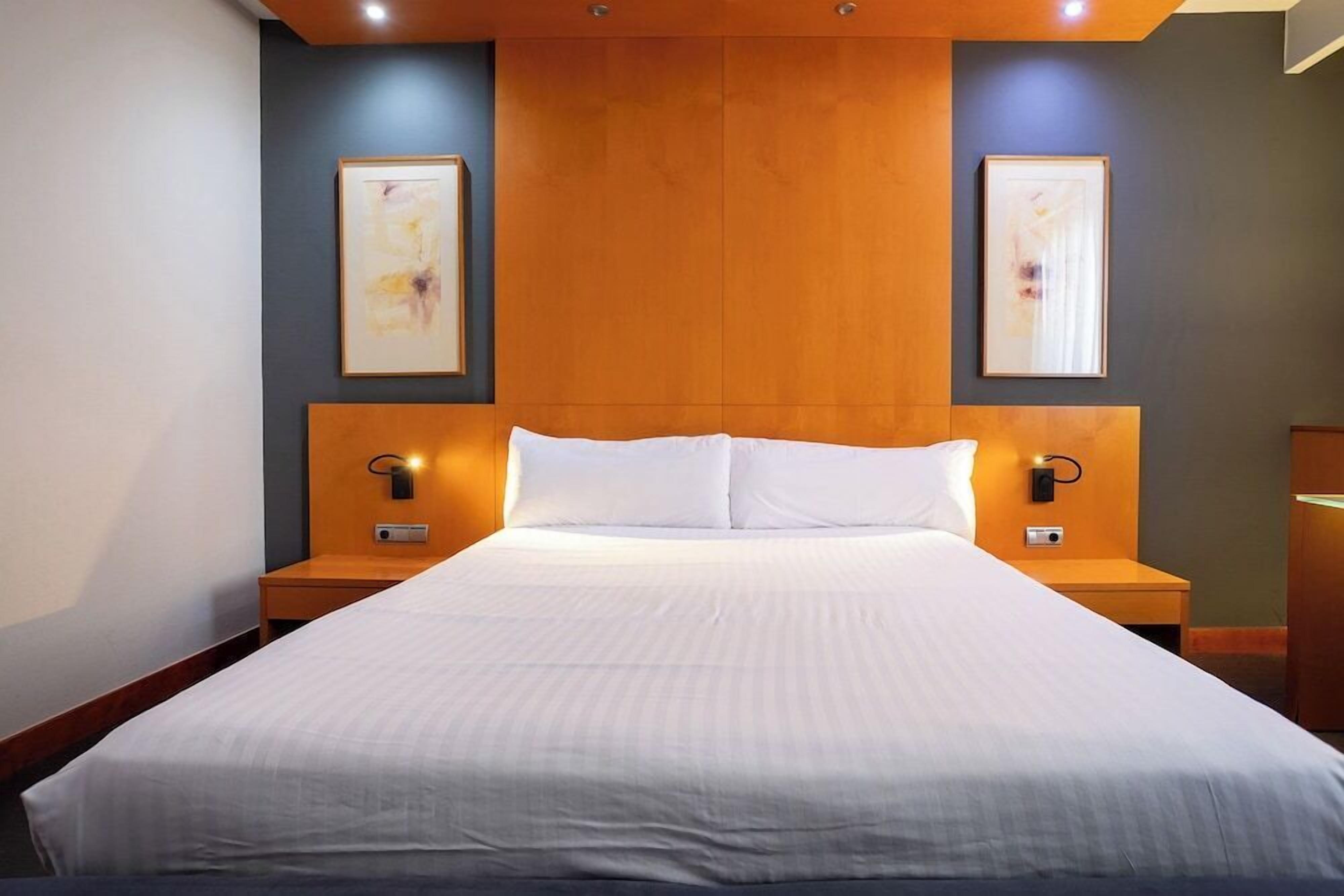 Een hotelkamer met een groot bed, een houten hoofdeinde, twee nachtkastjes en kunstwerken in lijsten aan de muren.