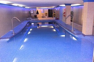 Una amplia piscina cubierta con suelo y paredes de azulejos, rodeada por una terraza con sillas de descanso y una zona de estar pequeña con una mesa y sillas.