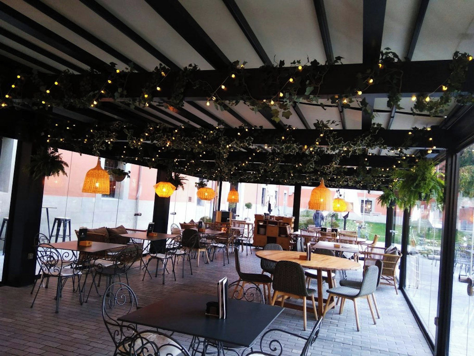 Un café al aire libre con mesas y sillas, luces colgantes y plantas suspendidas.