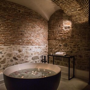 Un bagno con una vasca da bagno ampia, un muro in pietra e un piccolo tavolo con un asciugamano.