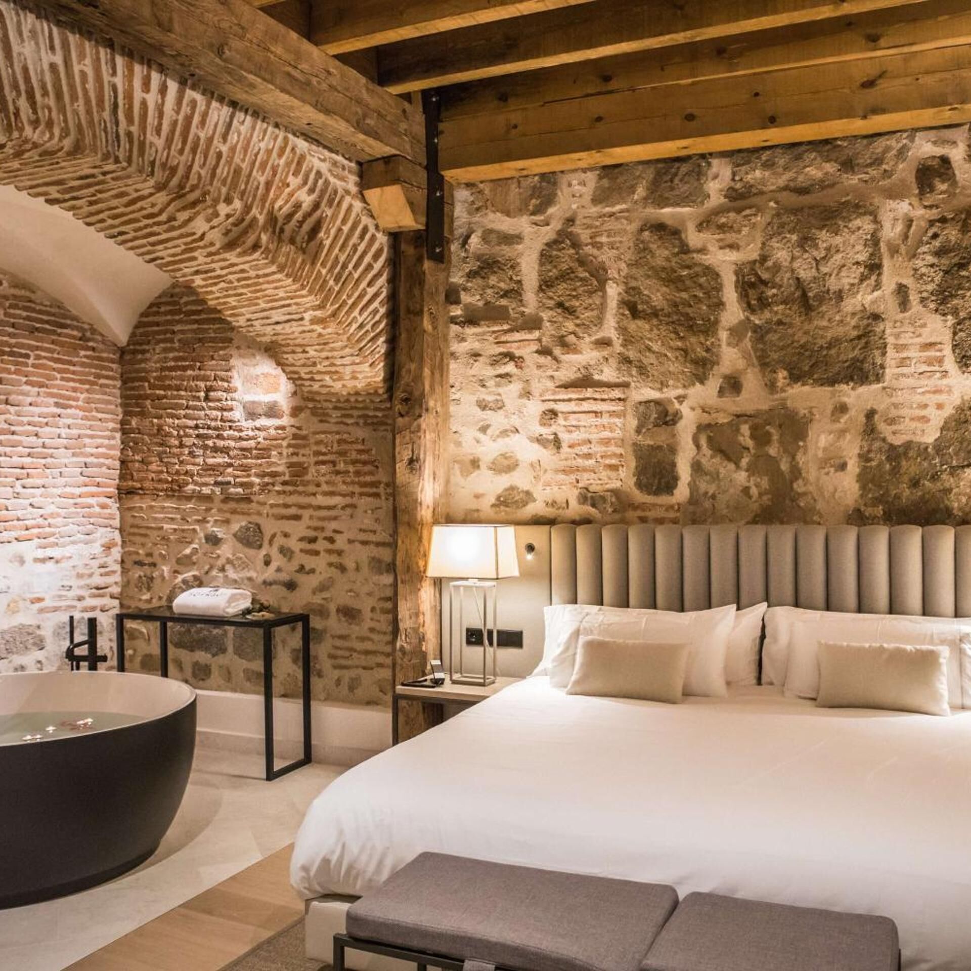 A room with a stone wall, a large bed, a black bathtub, and a wooden ceiling.