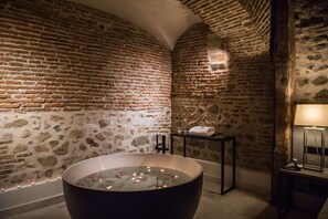 Un bagno con pareti in pietra, una vasca da bagno indipendente, un piccolo tavolo con un asciugamano e una lampada.