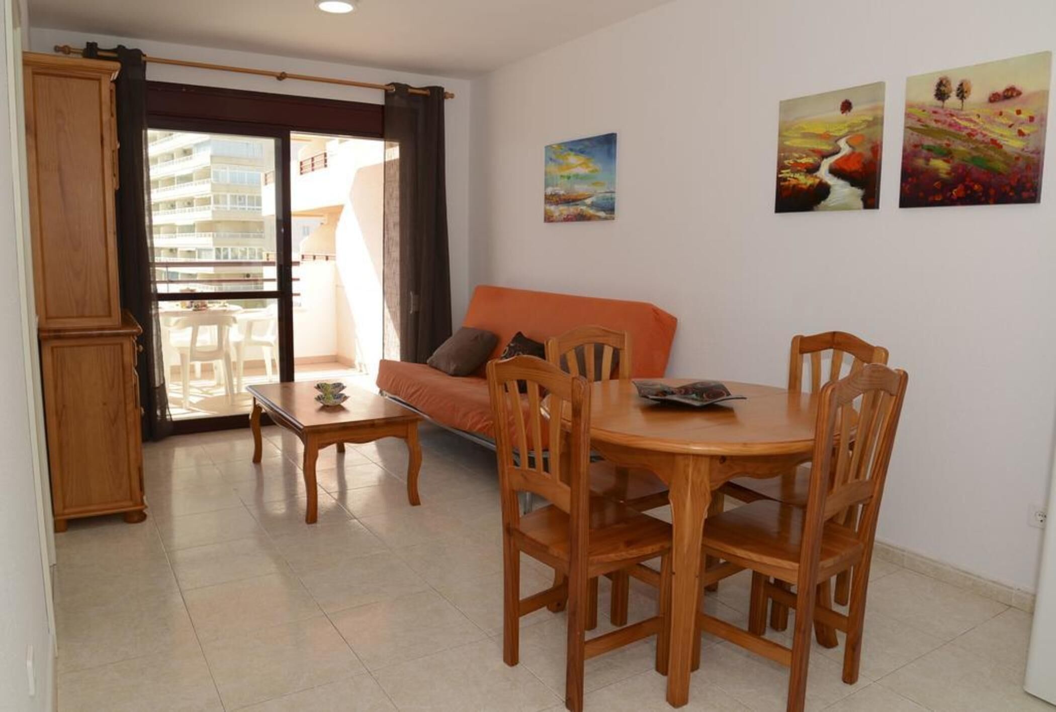 Zona de estar con sofá, mesa de comedor, sillas, un mueble de madera y un balcón con vistas a edificios.