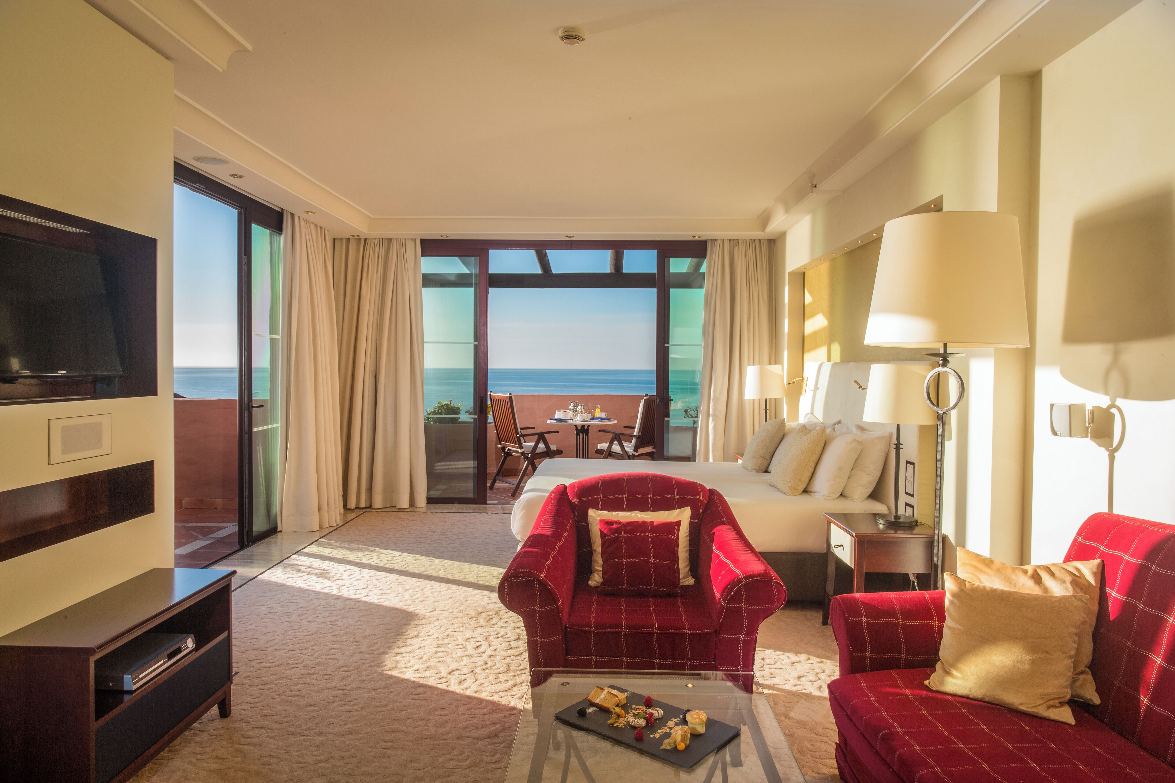 A hotel room with a large TV, a red armchair, a sofa, a dining table with chairs, a bed, and a balcony with a view of the sea.