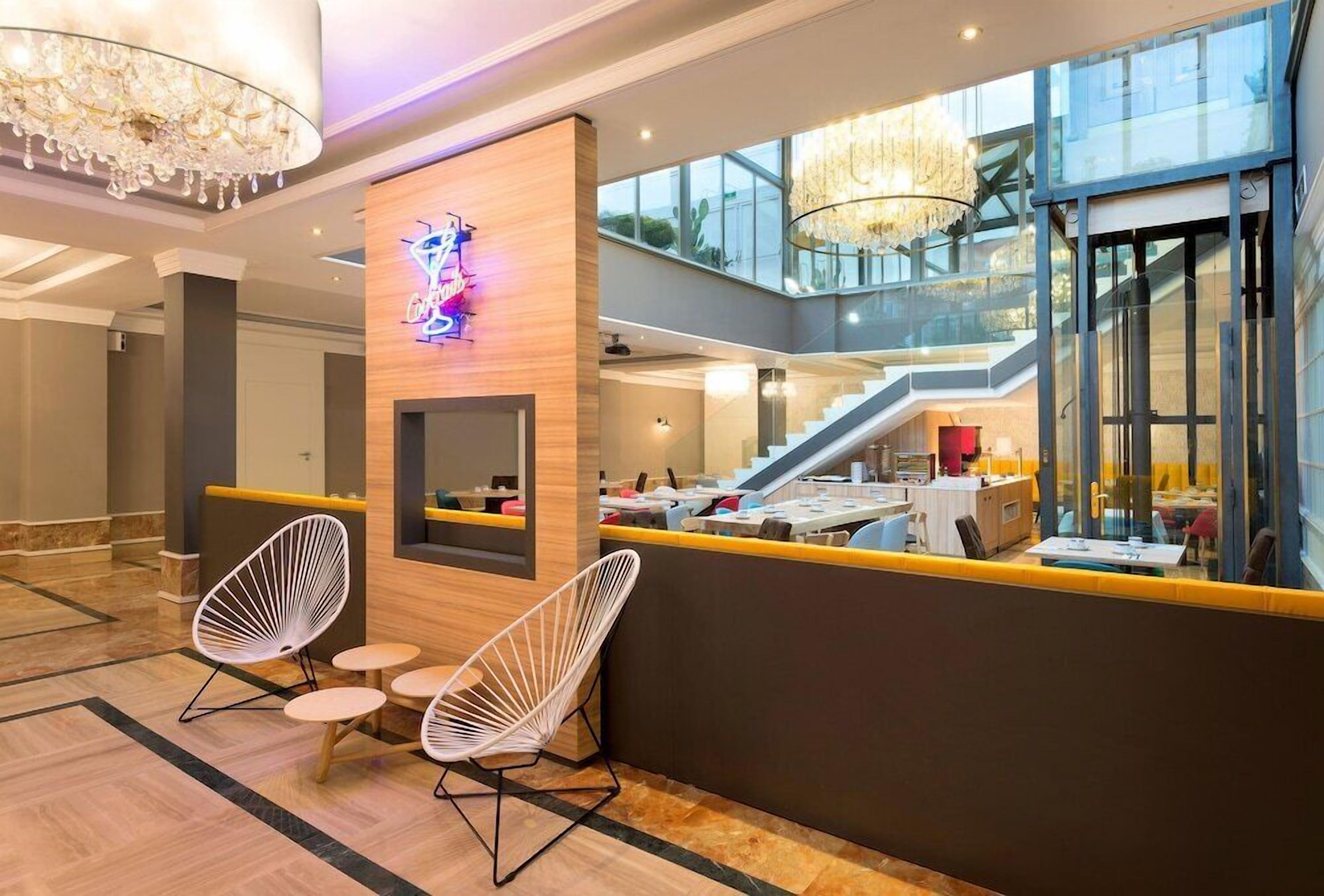Un vestíbulo de hotel moderno con un mostrador de recepción, dos sillas blancas de estructura metálica, una araña y una escalera que sube a un nivel superior.