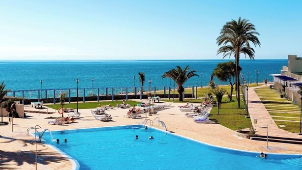 A pool area with a view of the sea, palm trees, and lounge chairs. - Grupotel Playa Feliz (San Bartolomé de Tirajana)