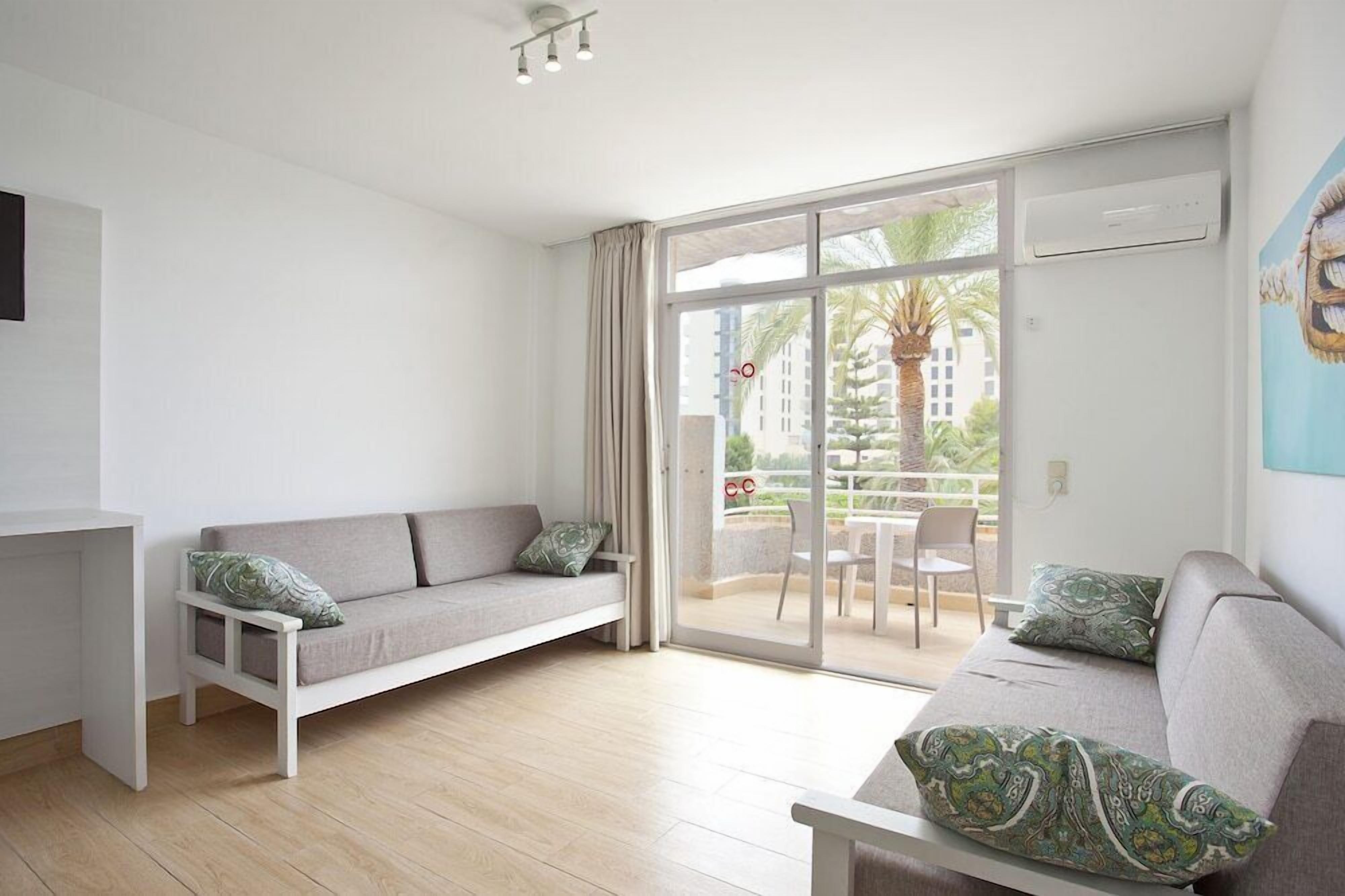 Una sala moderna con un sofá gris, un sillón blanco y una mesita. Hay una puerta corrediza de vidrio que da a un balcón exterior con vistas a palmeras y edificios.