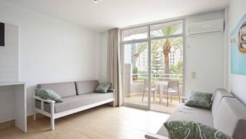 Una sala moderna con un sofá gris, un sillón blanco y una mesita. Hay una puerta corrediza de vidrio que da a un balcón exterior con vistas a palmeras y edificios.