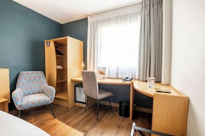 A compact hotel room with a desk, a chair, a wardrobe, and a window with curtains. - Travelodge Alicante Puerto (Alicante)
