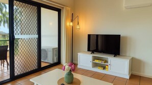 Een woonkamer met een televisie op een witte kast, een tafel met een bloevas en een glazen schuifdeur naar een buitenruimte.