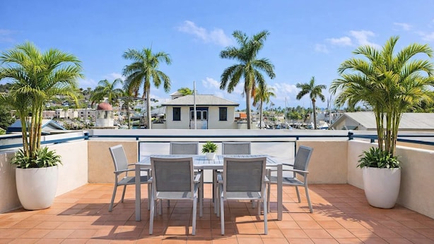 Een terras met een tafel en stoelen, omringd door plantenpotten, met uitzicht op een jachthaven met boten en een gebouw op de achtergrond.